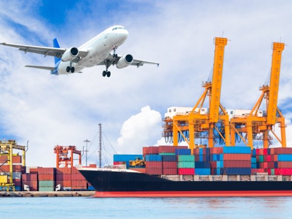 The plane is flying and a cargo is ship anchoring in ocean port.